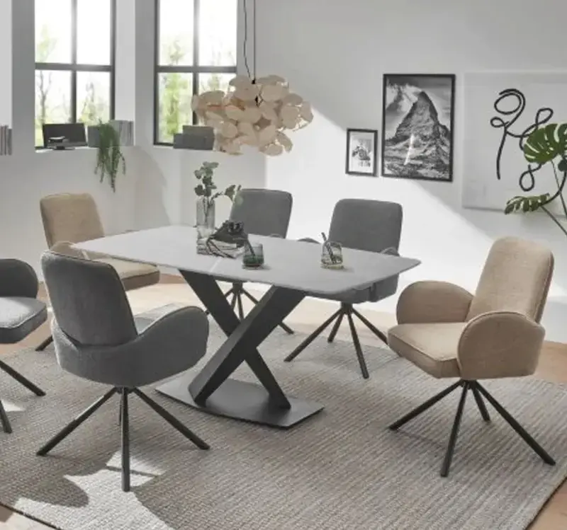 Essbereich mit rechteckigem Tisch auf dunkelgrauem Kreuzgestell, grauen und beigen Polsterstühlen auf Teppich, hinten Fensterbank mit Pflanzen und gerahmten Bildern.